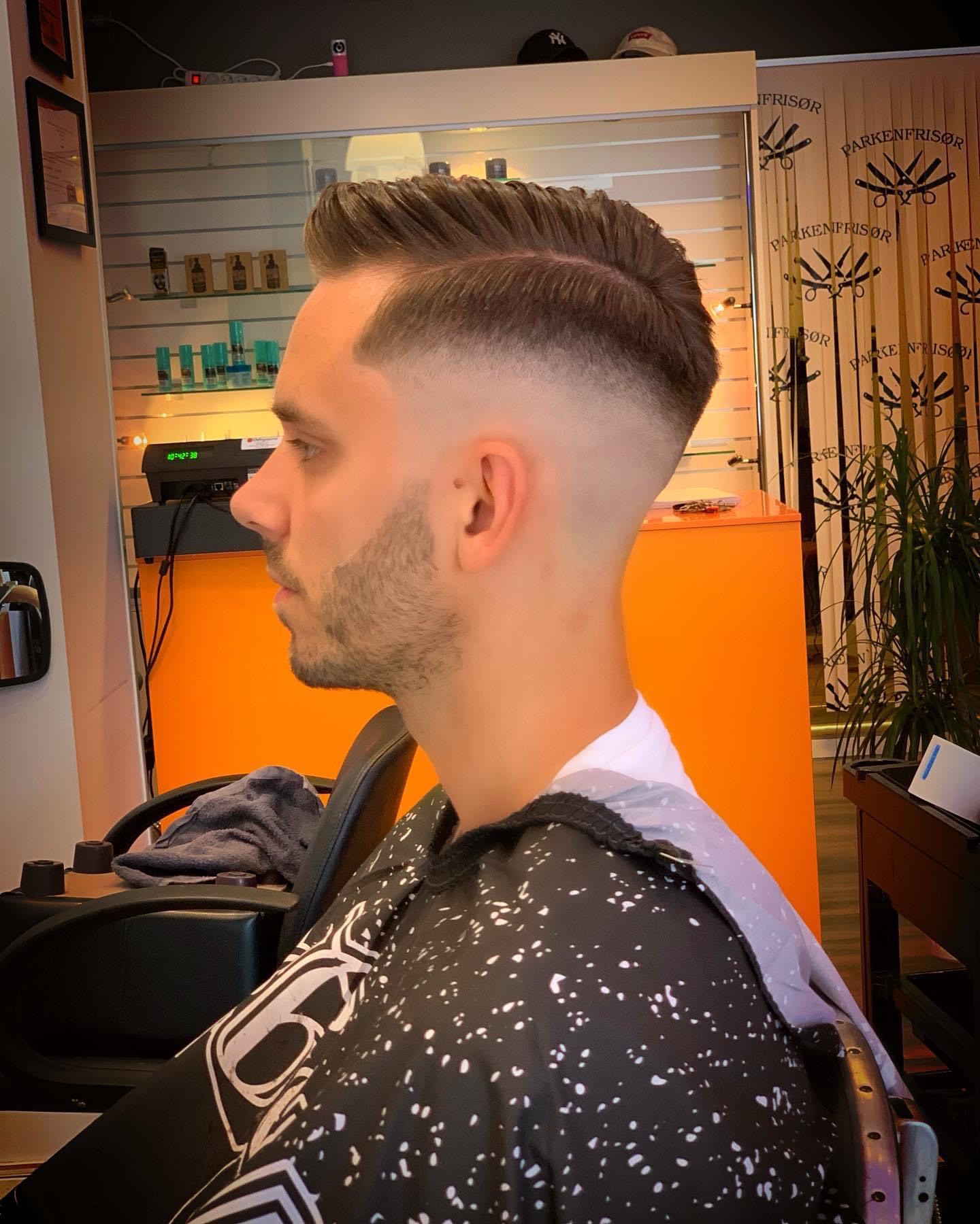 En mann med en skarp og presis skinnfade-frisyre. Overgangen fra hud til hår er sømløs og jevn, og gir et moderne og stilrent uttrykk. Perfekt for en velstelt og fresh look.